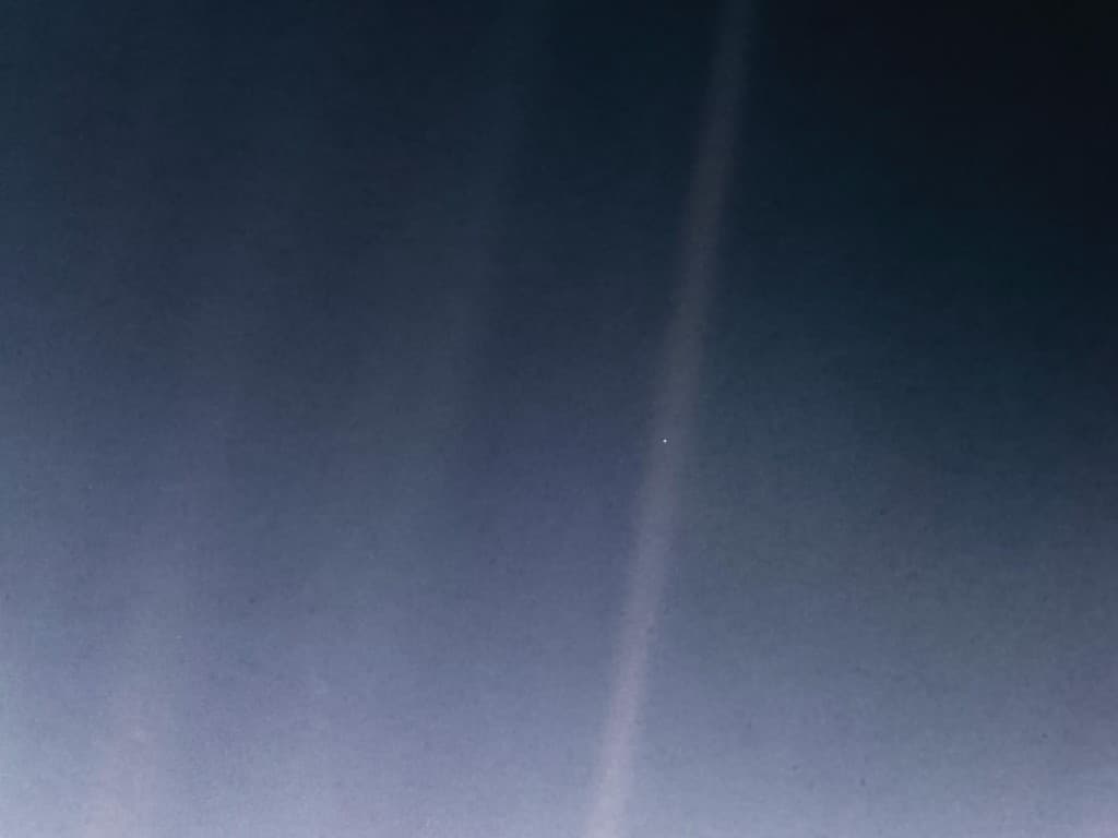 Voyager 1’s photograph of Earth: a single pale point of light in a band of sunlight, against the dark of space.